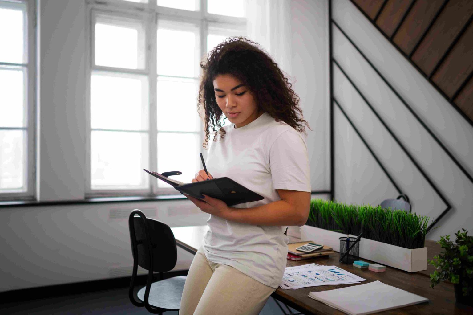 Woman writing in a notebook while sitting on a desk in a bright office.
