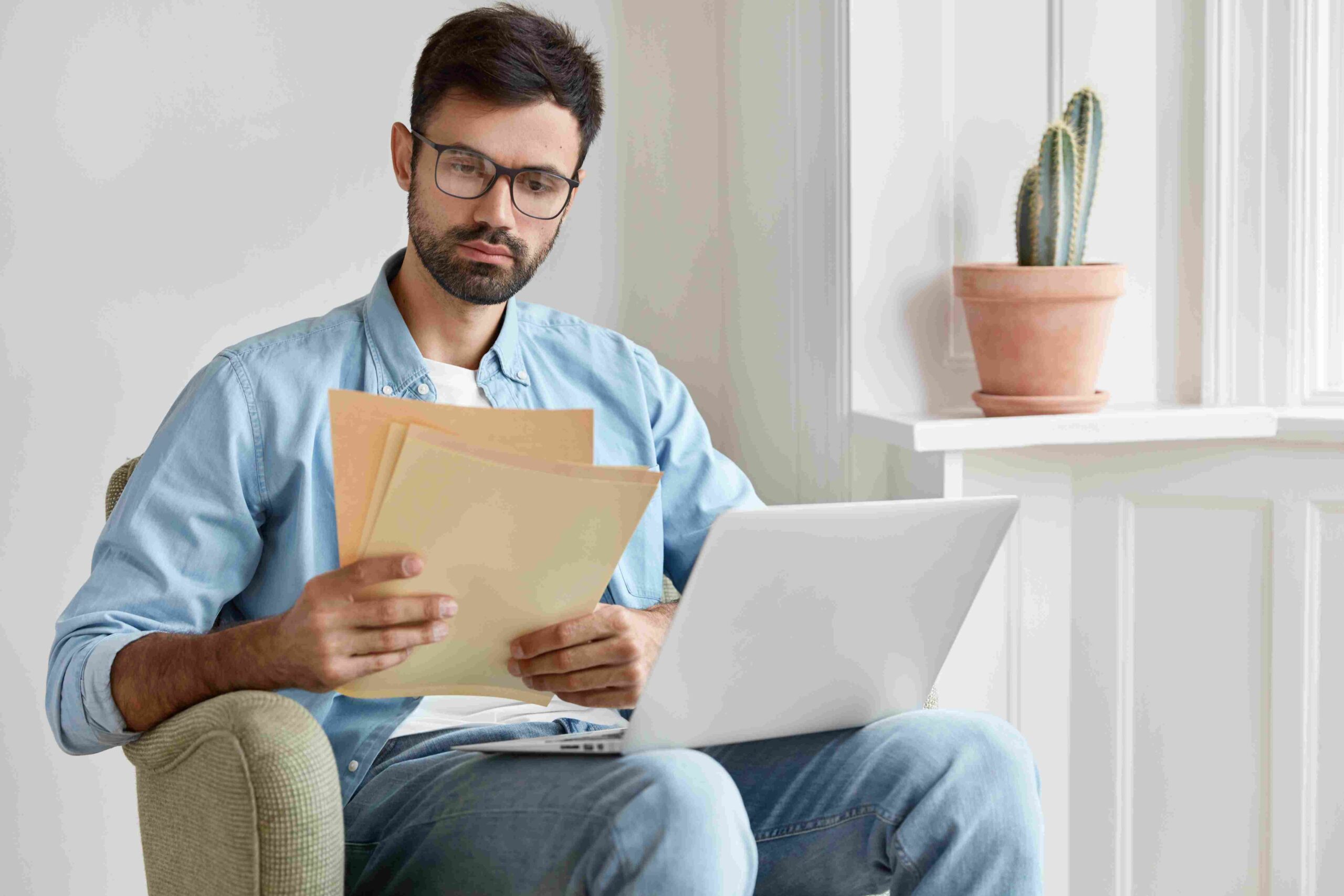 Man sitting at home reviewing documents while using a laptop, with a cactus on the windowsill in the background.